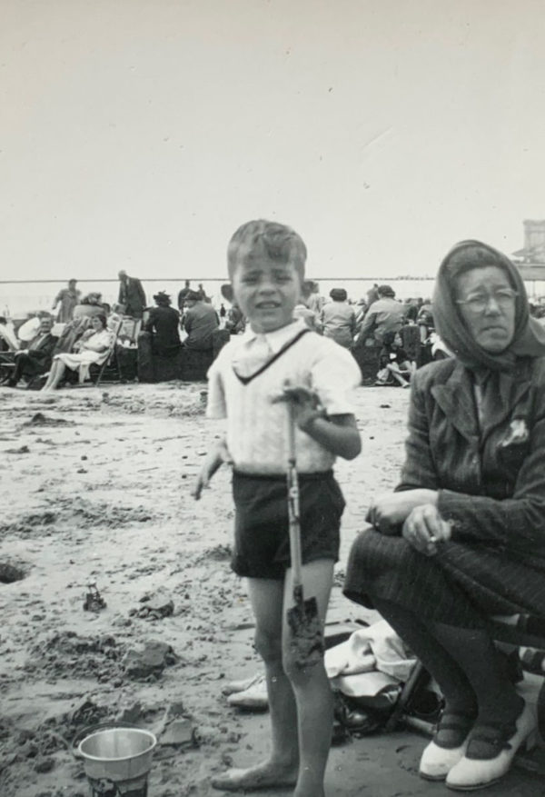 A black and white photo of a young Bobby standing on a beach alongside his mother, who is sat to his right,