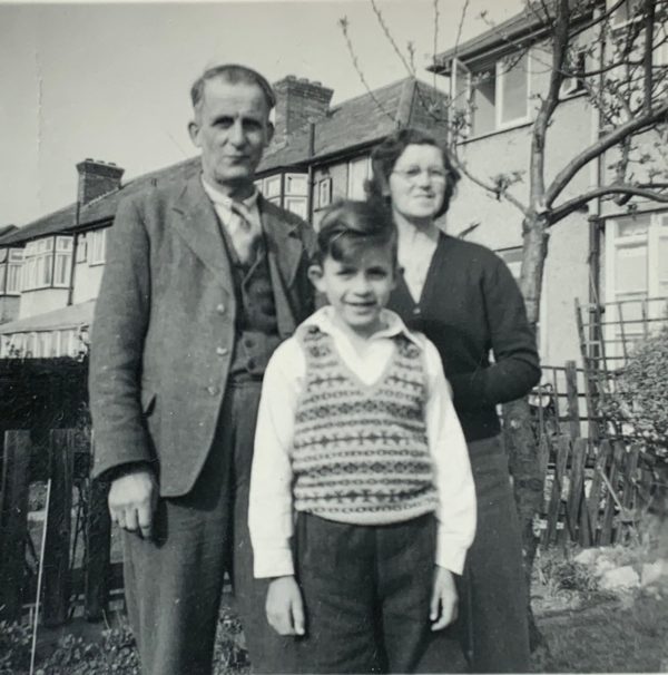 An older Bobby stood in front of his parents in the back garden of their house. A sense of belonging?