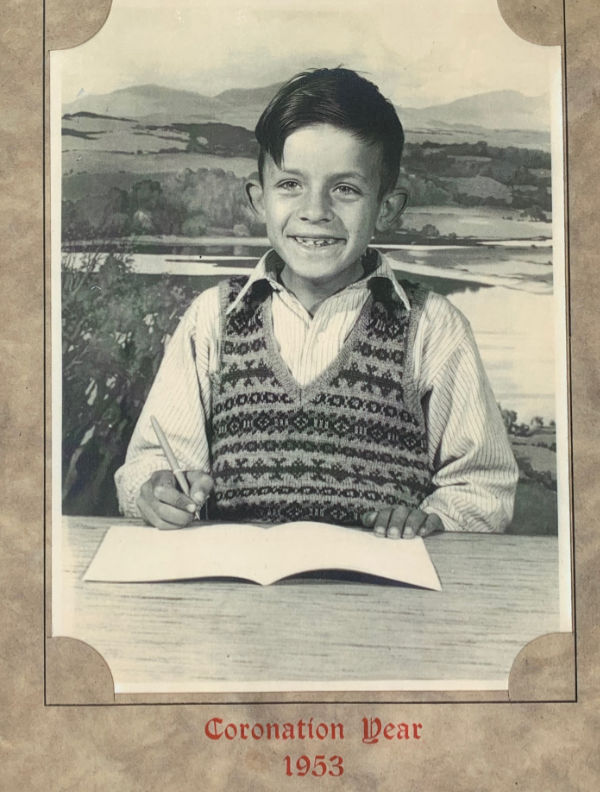 Bobby posing for a photograph at a desk with an open excercise book and pen in his right hand. He is wearing a Fair Isle jumper knitted by his Mum. The photo is in a frame , captioned Coronation Year 1953.