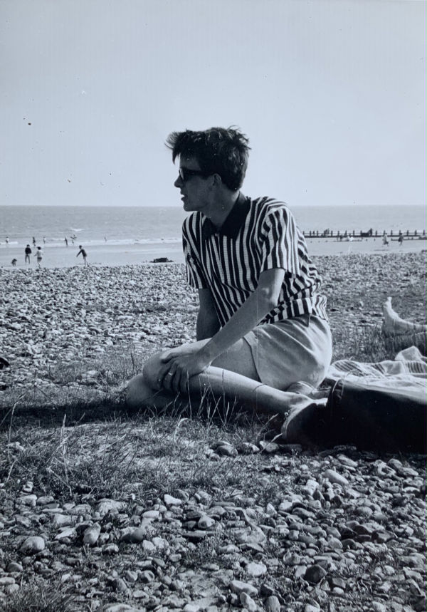 Bobby sat on a pebble beach. He is looking over the expanse of pebbles towards people playing in the sea. There is some grass growing through where he is sat.