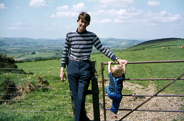 Bobby posing by a gate facing the camera, whilst his son is climbing up on the gate. The rolling hills of the Purbeck peninsula are in the background.