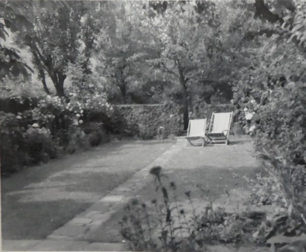 Black and white shot of the garden. Laid mainly to a well-cut lawn, with two deck chairs at the far end. A hedge surrounds the garden.