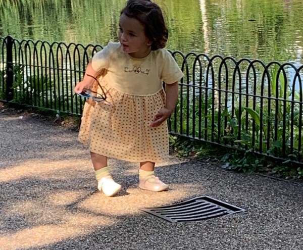 Picture of a young girl, holding a pair of glasses in her right hand, looking at a drain cover in the path.