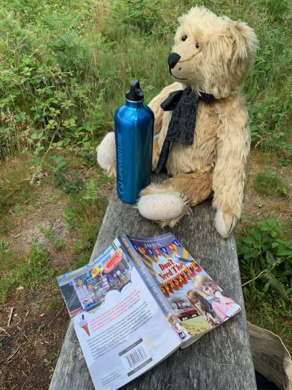 Close up of Bertie on Diddley's Bench. In front of him is a thermal water bottle, to his side an open book, face down, entitled "Don't need the Sunshine" by John Osborne.