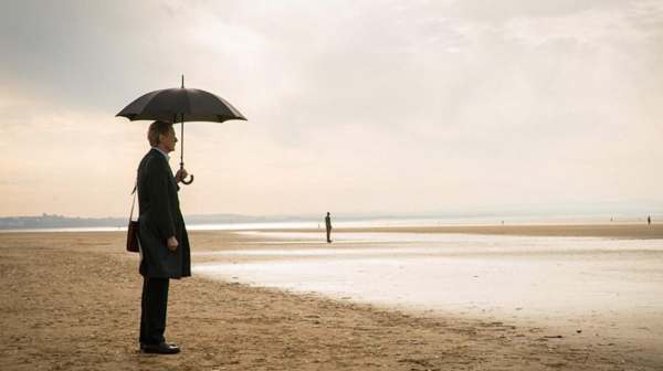 Picture of a beach on a grey day. Several loan people standing dotted around in the distance. In the foreground, a man in a coat, satchel over his left shoulder, holding an umbrella in his left hand.