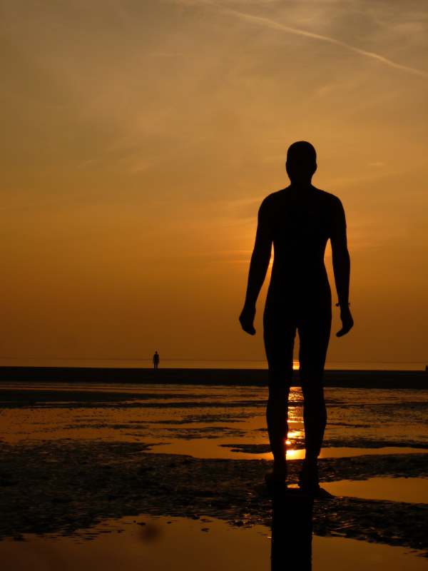 Looking at the back of a Gormley statue on Crosby Beach "Another Place" against the setting sun. The sky is a deep orange colour and the setting sun is reflecting on the water between the statue's legs.