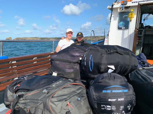 The two Sams posing on the boat behind the pile of luggage. Skomer is visible behind them.