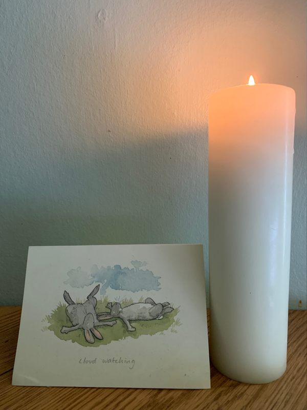 A candle lit for Diddley alongside a beautiful art water colour card of two hares lying on their backs cloudwatching.