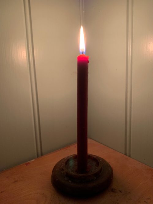 A lit candle on a table in a corner. For Diddly and the Haemophilia Society.