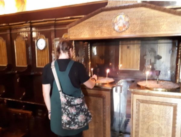 Lighting a Candle for Diddley: Emma, a lady with pig tails, stands with her back to the camera lighting a candle on an ornate table in front of her.