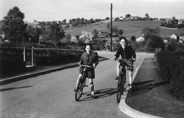 A boy (Chris), and a girl (his sister Elizabeth) in Brownie Uniform, both on bikes, but stopped at the top of a short hill. Chris is on the ex Paratrooper's Bicycle.