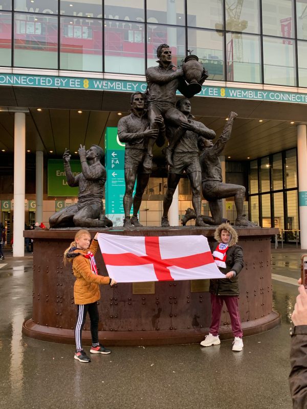 Two girls holding an England flag in front of a Rugby League statue.