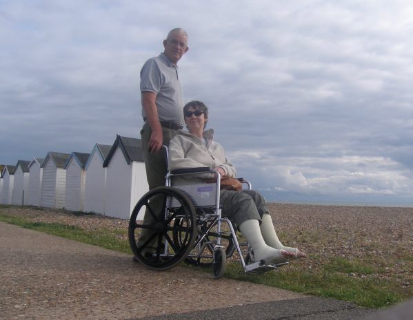 Bobby pushing Diddley (who has both ankles in plaster) in a wheelchair along Goring-by-Sea seafront, behind som ewhite beach huts.