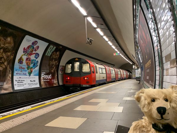 Platform at Colliers Wood. Bertie is sitting on a bench as an Underground train departs away from the camera.