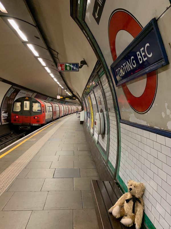 Bertie sat on a bench at Tooting Bec Station. A roundel is above the bench. A train is departing, away from the camera, heading to Morden.