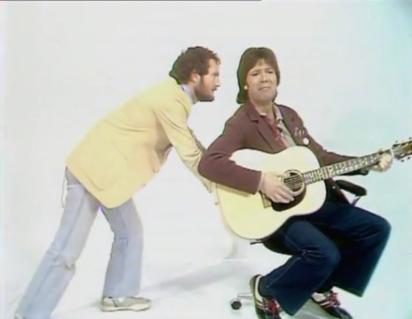 Cliff Richard playing guitar sitting on a typists chaier, with Kenny Everett wheeling him away...