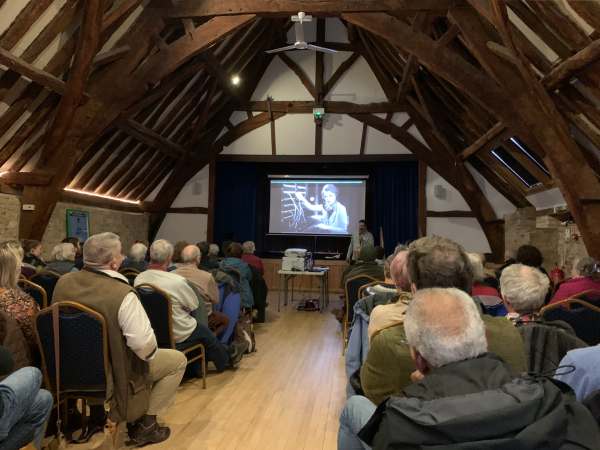 Interior of the Tithe Barn at Bishop's Cleeve. A polished wooden floor underneath an arched roof. A video presentation is being played on a screen on the front wall.