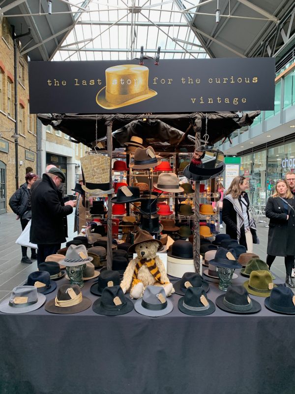 Bertie, wearing his Sutton United scarf and a hat, sat on the vintage hat stall in Spitalsfield Market.