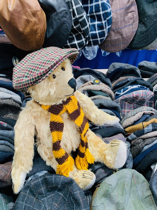 Close up of Bertie, wearing his Sutton United scarf and a flat cap, sat amongst the flat caps on the vintage hat stall in Spitalfields Market.