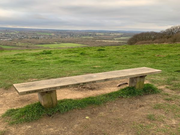 Plain bench overlooking the North Downs, with the inscription: Pilgrims come wind come weather. Dedicated to …