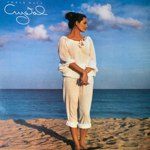 Album cover - "These Days", Crystal Gayle. Photograph of the artist in a white summer top and trousers on a beach,