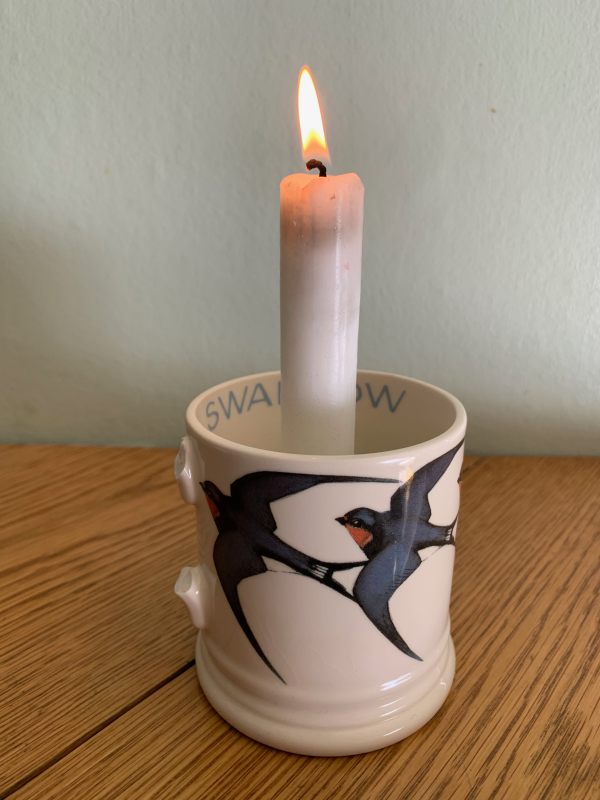 A candle lit for Diddley in a mug with painted swallows on it. The handle is broken.