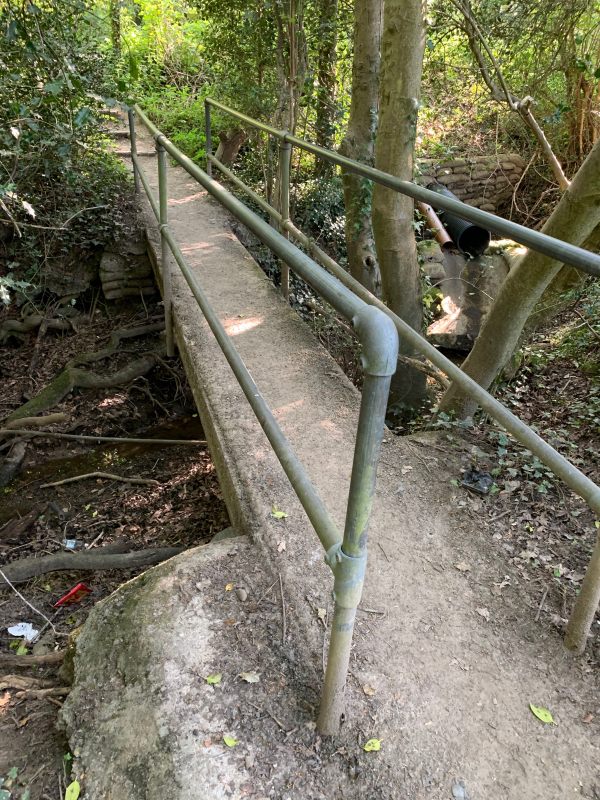 A simple narrow footbridge made of a substantial plank of wood with hand rails made from scaffold poles.