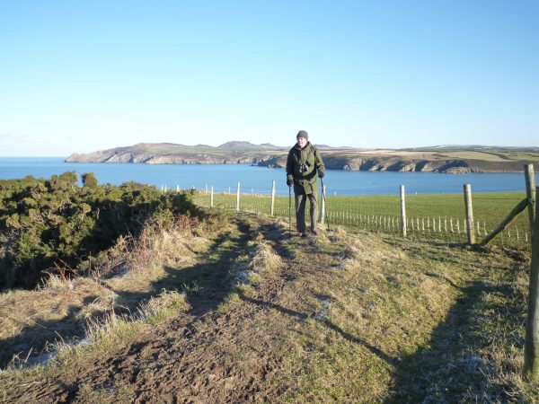 Bobby on the Pembrokeshire Coast Path, with the sea behind. He is wearing a green wooly hat and scarf, warm coat and has his hiking sticks in his hands.