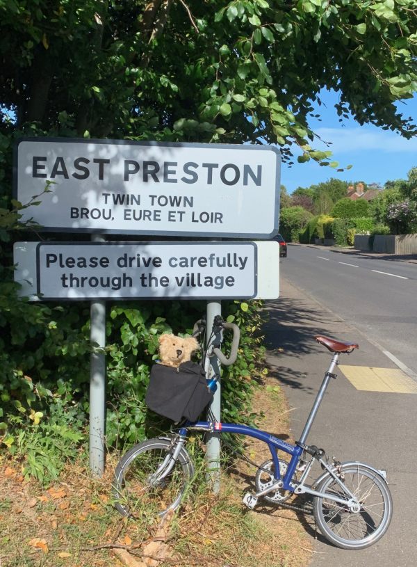 Bertie in the front basket of Bobby's bicycle leant up against the village sign for East Preston.