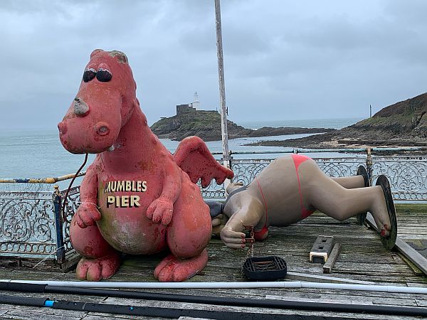 A dragon model with "Mumbles Pier" on its chest, and a large bikini-clad model lady, face down on the pier.