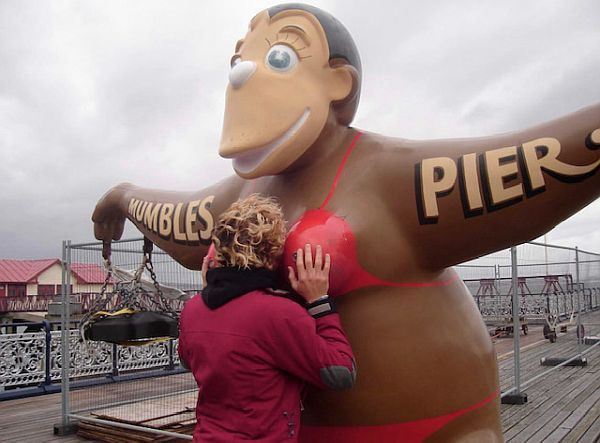 A bikini-clad monkey with "Mumbles Pier" across its arms. A lady has her face buried in its cleavage!