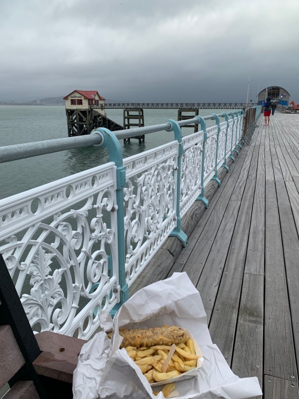 Looking down Mumbles Pier, with Bobby's Sausage & Chips in paper on a bench in the foreground.