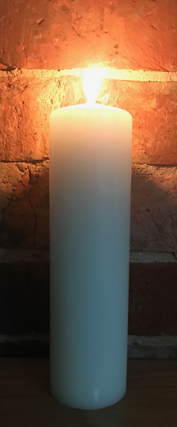 A tall, white candle lit for Diddley (and Gerry Marsden) against a plain brick wall.