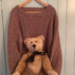 Bertie sat on Wendy's Jumper, knitted brown with cable patterning, on a hanger. The sleeves are wrapped around Bertie.