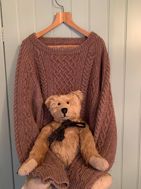 Bertie sat on Wendy's Jumper, knitted brown with cable patterning, on a hanger. The sleeves are wrapped around Bertie.