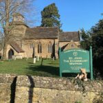 Bertie sat on a wall in front of the sign for Wotton Church, with the church in the background.