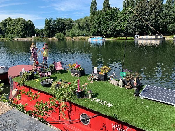 Narrow boat with synthetic grass on the roof along with flower-filled planters.